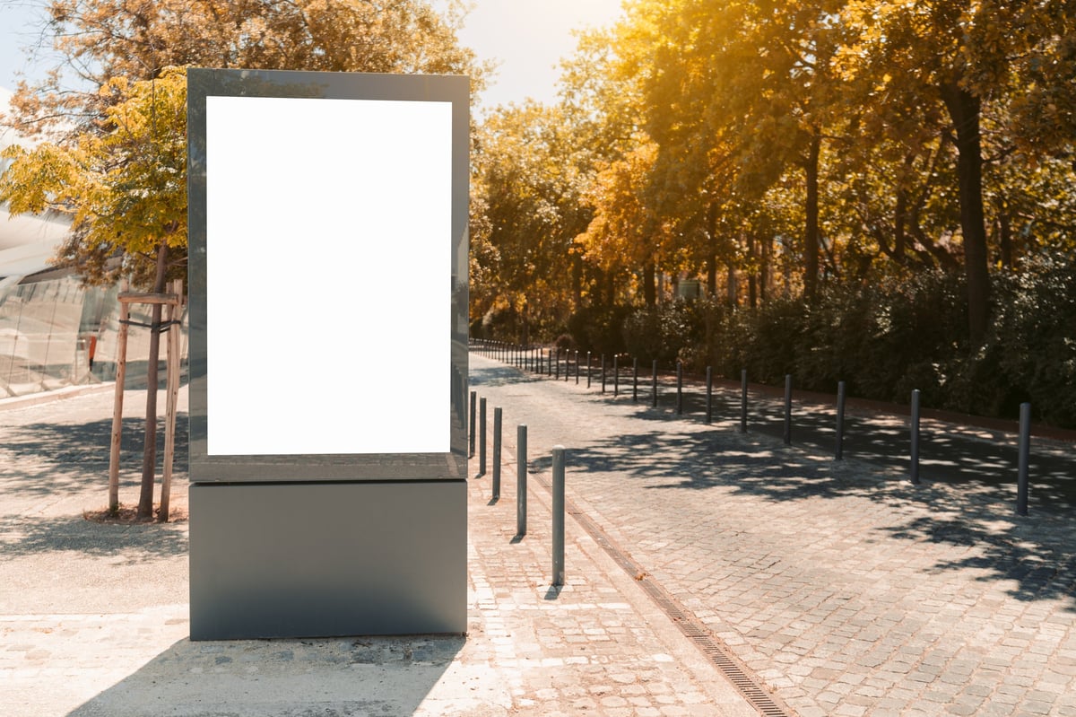 Large blank vertical digital street display beside a paved road and cycle lane under sunlit trees (stock photograph)
