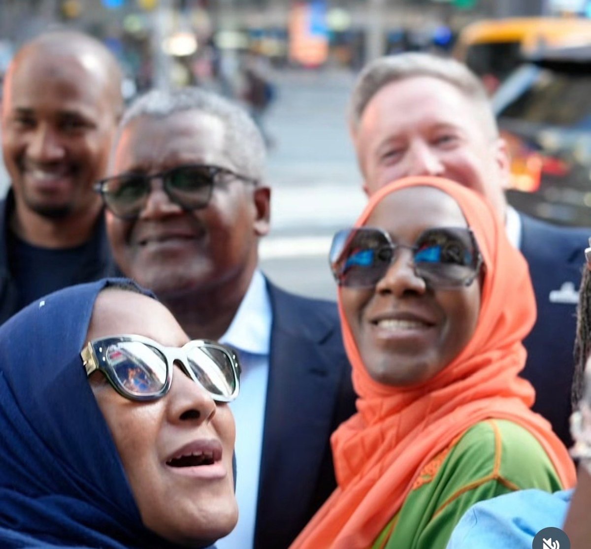 Group of five people smiling at camera outdoors, wearing sunglasses and casual clothing with urban street background
